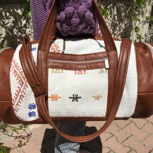 Puede incluir: Una bolsa de viaje blanca y marr&oacute;n con un bolsillo con cremallera y detalles bordados. La bolsa tiene detalles y correas de cuero marr&oacute;n.