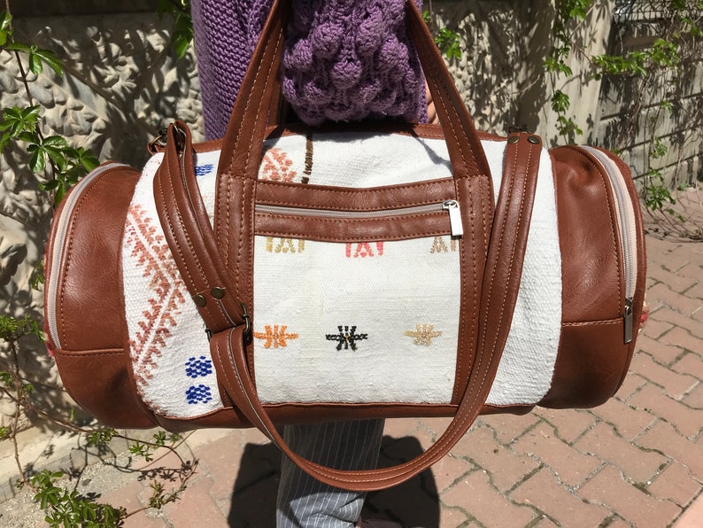 Puede incluir: Una bolsa de viaje blanca y marr&oacute;n con un bolsillo con cremallera y detalles bordados. La bolsa tiene detalles y correas de cuero marr&oacute;n.