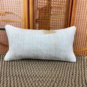 Pode incluir: Uma almofada lombar retangular com uma capa de tecido texturizado em bege claro. A almofada tem um design simples e limpo com um pequeno detalhe de remendo castanho de bordas irregulares centrado perto da borda superior, criando um contraste.