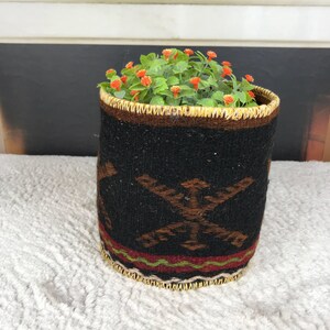 May include: A cylindrical woven fabric planter featuring a black base with brown geometric patterns and a red and green horizontal stripe near the bottom. The top and bottom edges are finished with a light tan braided trim, holding a green plant.