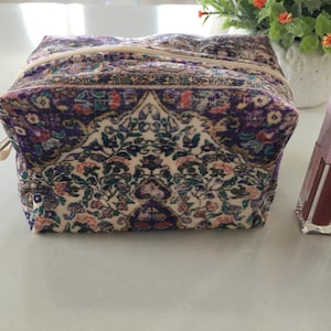 Pode incluir: Uma bolsa de cosméticos quadrada com um padrão floral em tons de roxo, creme e verde. A bolsa tem um zíper dourado e está ao lado de um tubo de batom e um frasco de esmalte. A bolsa tem aproximadamente 15 cm de largura.
