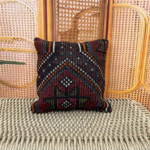 Puede incluir: Cojín decorativo cuadrado con un patrón geométrico en tonos burdeos, azul marino, naranja y verde. El cojín está colocado sobre una superficie tejida beige, con un fondo de madera natural.