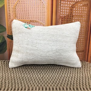 Op de afbeelding: Een rechthoekig, crèmekleurig kussen met een getextureerd oppervlak. Een klein, kleurrijk bloemmotief is zichtbaar in de bovenhoek. Het kussen is geplaatst op een geweven, bruin oppervlak, wat duidt op een woondecoratie-artikel.