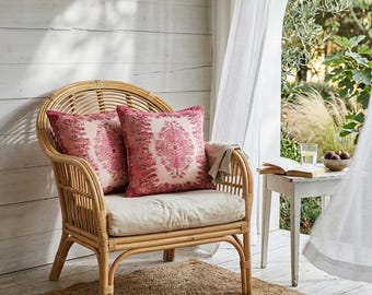 Puede incluir: Un sillón de ratán natural con dos cojines decorativos rosas. Los cojines presentan un detallado estampado floral. La silla está sobre una alfombra de yute tejida, con una mesita auxiliar blanca y cortinas transparentes en el fondo.