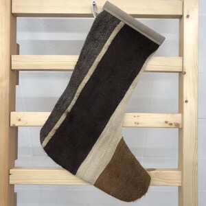 Puede incluir: Una media de color marrón, blanco y beige con rayas y textura tejida. La media está colgada de una escalera de madera.