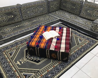 May include: A grey and black patterned floor rug with a colourful patterned square ottoman in the centre. A book is resting on top of the ottoman.