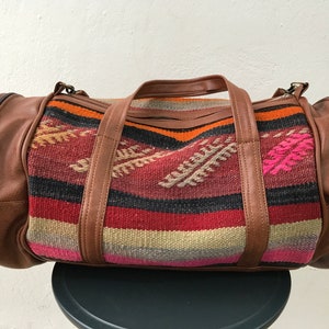 Op de afbeelding: Een bruine leren reistas met een kleurrijk geweven patroon. Het patroon heeft strepen in rood, oranje, roze, zwart en beige.