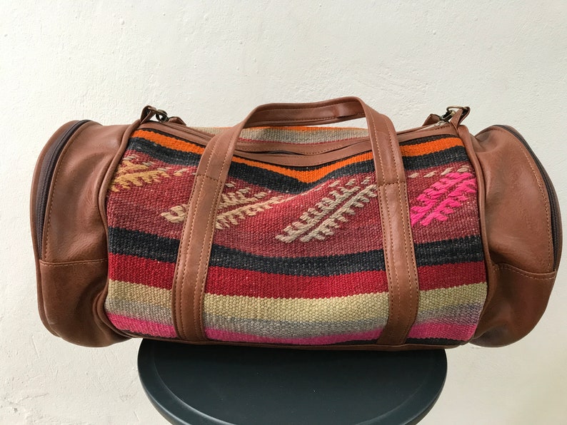 Op de afbeelding: Een bruine leren reistas met een kleurrijk geweven patroon. Het patroon heeft strepen in rood, oranje, roze, zwart en beige.