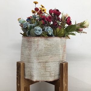 May include: A white and brown woven fabric planter with a variety of colorful artificial flowers, sitting on a wooden stand.