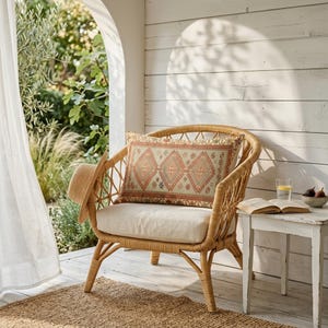 May include: A wicker chair with a patterned pillow, a straw hat, and a small side table. The pillow has a geometric design in shades of red, beige, and green. A book, a glass of water, and a bowl of figs are on the table.