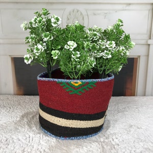 Puede incluir: Un macetero tejido de color rojo, negro y blanco con un diseño verde y amarillo. El macetero está lleno de una planta con flores blancas.