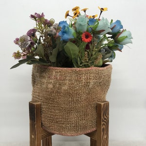 Puede incluir: Una cesta tejida marrón con un borde rosa llena de un ramo de flores artificiales, incluyendo rosas azules, margaritas amarillas y flores moradas. La cesta está sentada en un soporte de madera.