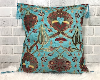 Puede incluir: Una almohada azul turquesa con un patrón floral turco tradicional en tonos rojos, verdes y dorados. La almohada tiene borlas en cada esquina.