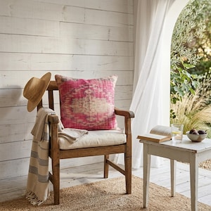 Può includere: Una sedia in legno con un cuscino a fantasia rosa e bianco, un cappello di paglia e una coperta a righe. Un tavolino bianco con un libro, un bicchiere d'acqua e una ciotola di fichi. La scena è su un tappeto di iuta.