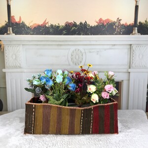 Può includere: Un cesto rettangolare intrecciato con un motivo a strisce marroni, rosse e gialle. Il cesto è pieno di una varietà di fiori artificiali, tra cui rose blu, rose rosa e fiori gialli.