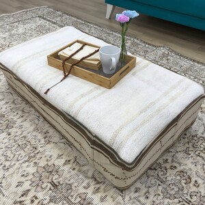 May include: A white and brown patterned ottoman with a wooden tray on top. The tray has a white mug, a string of wooden beads, and a small bouquet of pink flowers in a glass vase.