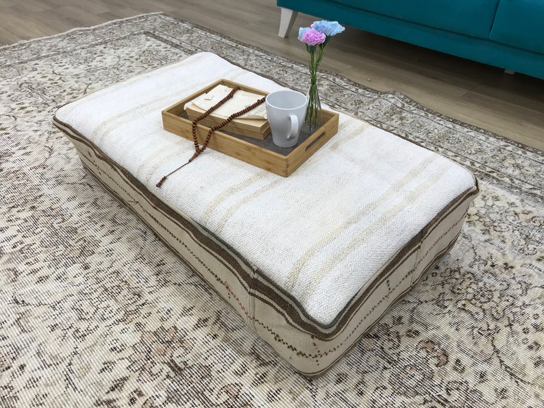 May include: A white and brown patterned ottoman with a wooden tray on top. The tray has a white mug, a string of wooden beads, and a small bouquet of pink flowers in a glass vase.