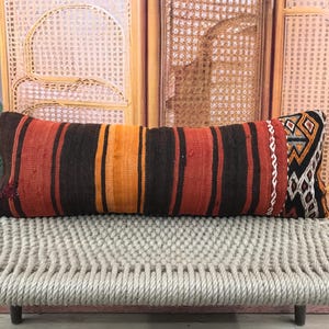 Peut inclure: Un long coussin rectangulaire avec un motif à rayures dans les tons marron, orange et rouge. Le coussin a une extrémité décorative avec des motifs géométriques noirs, blancs et orange. Il repose sur un banc tressé.