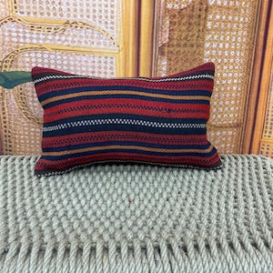 Puede incluir: Cojín rectangular con un estampado de rayas en rojo, azul marino y dorado. El cojín está colocado sobre una superficie tejida. El diseño presenta rayas horizontales, creando una apariencia texturizada. La combinación de colores es cálida y acogedora.