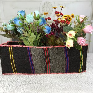 Op de afbeelding: Een geweven rechthoekige mand met een roze rand, gevuld met verschillende kunstbloemen, waaronder blauwe, roze en gele rozen, en rode en gele wilde bloemen.