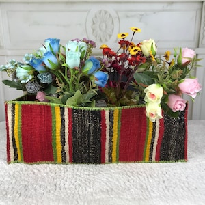 May include: A rectangular woven fabric basket featuring vertical stripes in red, black, yellow, and green. The basket is filled with an assortment of artificial flowers, including blue, pink, and cream-colored roses alongside small yellow and red blossoms.