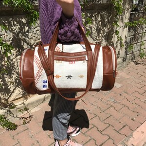 Puede incluir: Una bolsa de viaje blanca y marr&oacute;n con detalles bordados de colores. La bolsa tiene un ribete de cuero marr&oacute;n y un cierre de cremallera.