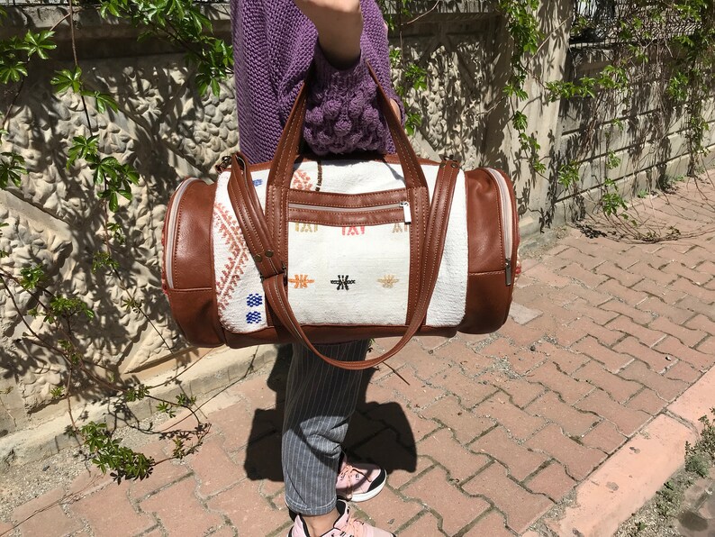 Puede incluir: Una bolsa de viaje blanca y marr&oacute;n con detalles bordados de colores. La bolsa tiene un ribete de cuero marr&oacute;n y un cierre de cremallera.