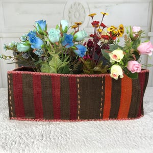 May include: A rectangular woven fabric basket featuring vertical stripes in shades of brown, dark red, and orange, with visible stitched edges. The basket is filled with an assortment of artificial flowers, including blue, pink, and yellow roses.