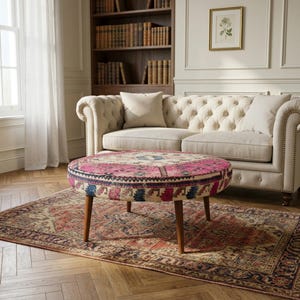 May include: A round ottoman featuring a vibrant, multi-coloured patterned fabric with prominent shades of pink, blue, and cream. The piece is supported by four tapered wooden legs and displays a detailed, intricate textile design across its surface.