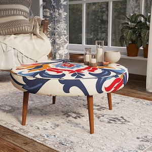 Op de afbeelding: Een ronde poef met een crèmekleurige basis en een opvallend, symmetrisch bloemmotief in marineblauw, rood en oranje. Het meubelstuk rust op vier taps toelopende houten poten en heeft een laag, cirkelvormig ontwerp.