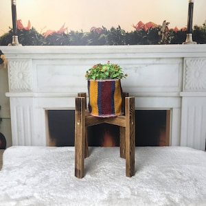 May include: A wooden plant stand with a potted plant in a colorful striped fabric planter. The plant stand is in front of a white marble fireplace with a garland of greenery on the mantle.