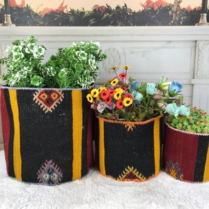 May include: Three cylindrical baskets made from woven fabric featuring a black, red, and yellow striped pattern with geometric diamond motifs. Each basket contains artificial greenery and flowers, including yellow, red, and light blue blossoms.