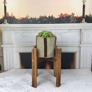 May include: A cylindrical woven fabric planter with vertical brown stripes and a light beige base, containing a small green plant with orange accents, resting on a dark brown wooden stand with four legs and a cross-shaped support structure.