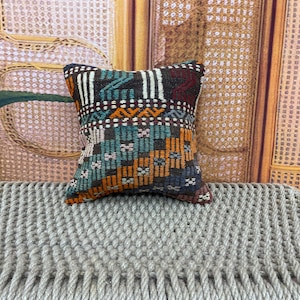 May include: A small, square decorative pillow with a geometric pattern. The pillow features a mix of colors, including teal, orange, brown, and white. The pillow is likely made of woven fabric and is placed on a textured surface.