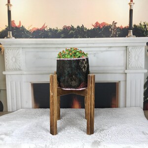 May include: A small potted plant with green leaves and orange flowers sits inside a dark, textured fabric container. This container is supported by a four-legged wooden stand featuring a dark brown, stained finish and a cross-brace design.
