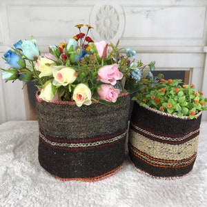 May include: Two woven baskets filled with colorful artificial flowers. The larger basket is dark brown with red and white stripes, while the smaller one has a beige stripe. The flowers include pink, blue, and yellow roses.