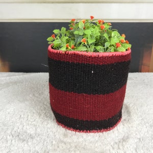 May include: A red and black striped woven planter with a green plant with small pink flowers inside.