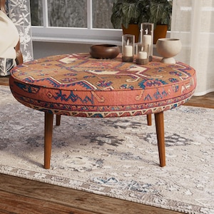 May include: A round ottoman with a low profile and four tapered wooden legs. The top is upholstered in a fabric featuring a multi-colored geometric pattern in shades of rust, blue, and cream, reminiscent of a traditional woven rug design.