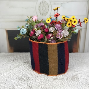 May include: A woven basket with red, blue, and brown stripes, filled with a bouquet of pink, yellow, and blue flowers.