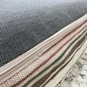 May include: Close-up of a grey and white striped cushion with a zipper closure. The cushion is made of a soft, woven fabric and has a subtle pink stripe.