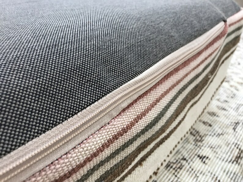 May include: Close-up of a grey and white striped cushion with a zipper closure. The cushion is made of a soft, woven fabric and has a subtle pink stripe.