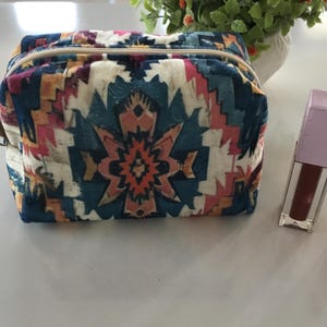 May include: A rectangular cosmetic bag with a colorful geometric pattern in teal, orange, pink, and cream. Next to it are two tubes of makeup: one with a brown liquid and a lavender cap, and another with a pink liquid.
