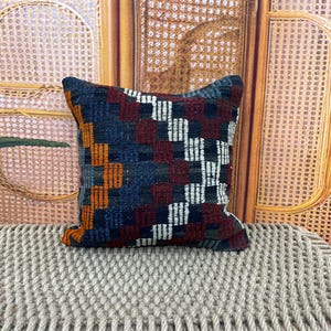 May include: Square decorative pillow with a woven geometric pattern in shades of blue, red, orange, and white. The pillow is placed on a textured, light-colored surface, with a woven background.