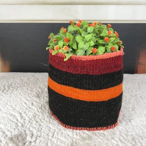 Puede incluir: Una maceta cilíndrica de tela tejida con rayas horizontales en negro, rojo oscuro y naranja brillante. Los bordes superior e inferior están rematados con una puntada de festón decorativa de color naranja. Un grupo de follaje verde con pequeñas flores naranjas.