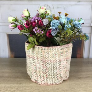 Pode incluir: Um cesto tecido cilíndrico com uma base de cor creme com subtis manchas cor-de-rosa e verdes, cheio de um arranjo de flores artificiais em tons de rosa, branco, roxo e azul claro, incluindo rosas e pequenas flores semelhantes a cravos.