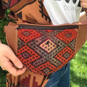 May include: A brown leather fanny pack with a zipper closure and a colorful woven fabric front panel featuring a geometric pattern. The fabric is in shades of red, orange, green, and brown.