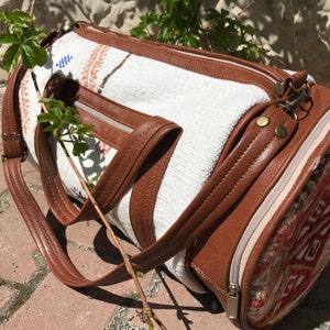 Puede incluir: Una bolsa de viaje cil&iacute;ndrica blanca y marr&oacute;n con cierre de cremallera y una correa de cuero. La bolsa tiene un patr&oacute;n tejido y un ribete de cuero marr&oacute;n.