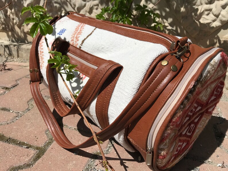 Puede incluir: Una bolsa de viaje cil&iacute;ndrica blanca y marr&oacute;n con cierre de cremallera y una correa de cuero. La bolsa tiene un patr&oacute;n tejido y un ribete de cuero marr&oacute;n.