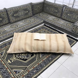 May include: A gray and black patterned floor cushion with a book on top. The cushion is on a gray and black patterned rug. The rug is on a white tiled floor.