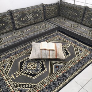 May include: A gray and black patterned floor cushion set with a matching rug. The cushions are arranged in a traditional L-shape and feature intricate geometric designs. A book is placed on a white pillow in the center of the rug.